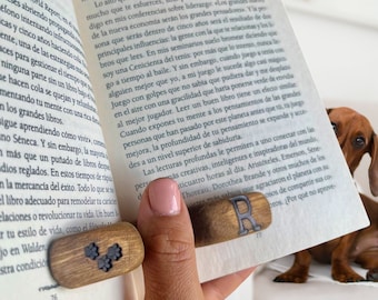 May include: A wooden bookmark with a metal letter 'R' on it. The bookmark is brown and has a natural wood grain. The bookmark is being held by a hand, and the tip of the bookmark is resting on a page of a book. The book is open and the page is filled with text. The text on the page is in Spanish.