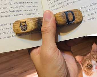 May include: A wooden bookmark with a cat design and the letter "D" burned into the wood. The bookmark is being held by a hand over an open book.