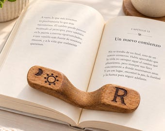 May include: A curved wooden book page holder resting on an open book. The left side features a laser-engraved crescent moon and sun icon, while the right side displays a capital letter R. The wood has a natural grain and a smooth, polished finish.