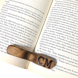 May include: A wooden bookmark with the initials "CM" engraved in black lettering. The bookmark is resting on an open book with text about a man named Chris.