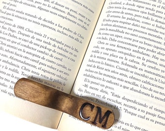 May include: A wooden bookmark with the initials "CM" engraved in black lettering. The bookmark is resting on an open book with text about a man named Chris.