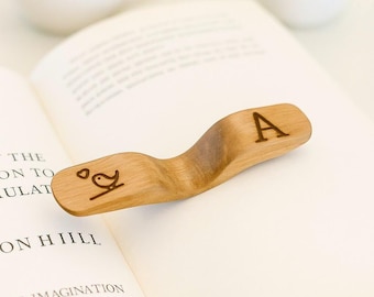 May include: Wooden bookmark with a curved design, featuring an engraved letter "A" and a small bird illustration. The bookmark rests on an open book, with a cup of coffee and a small potted plant in the background.
