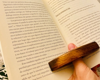 May include: A close-up of an open book with a wooden page holder. The book is open to a page with the title "Evita la trampa del 'lenguaje de victima'" and text about overcoming victim mentality. The page holder is made of dark wood and has a natural finish.