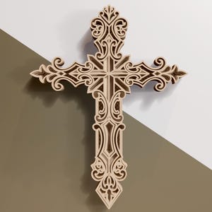 May include: A layered wooden cross with intricate, swirling cut-out designs. The cross is a light beige color and features a central starburst design. The background is a two-tone of white and a muted brown.
