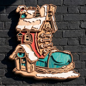 May include: A multi-layered wooden wall art piece shaped like a boot house. The design features a teal toe, a red midsection, and a snow-covered roof with pine trees. The structure includes a small attached cabin, windows, and lacing details.