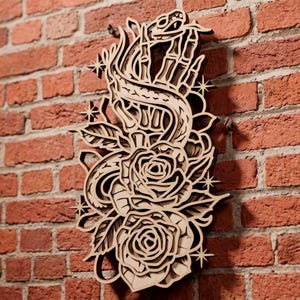 Puede incluir: Una pieza de arte mural de madera en capas que presenta una mano esquelética que se extiende hacia arriba, entrelazada con una serpiente y dos rosas en flor. El diseño incluye varios detalles de estrellas de cuatro puntas, todo elaborado con un acabado de madera clara natural y bordes oscuros.