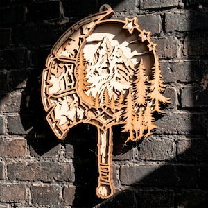 May include: A wooden wall hanging with a layered design depicting a mountain landscape, trees, and stars. The natural wood tones create a rustic aesthetic. The artwork is shaped like an axe, with intricate details and a decorative handle. It's mounted on a dark brick wall.