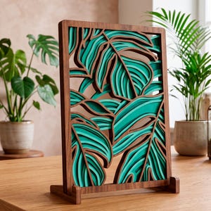 Puede incluir: Panel decorativo de madera con un diseño en capas de hojas tropicales en tonos turquesa y marrón. El panel, con marco y base de madera oscura, se exhibe sobre una mesa de madera. Una obra de arte para una decoración inspirada en la naturaleza.