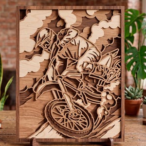 Op de afbeelding: Een gelaagd houten kunstwerk dat een motorcrosser in actie toont. Het stuk heeft een gedetailleerd ontwerp van de rijder en motorfiets, met wolken en terreinelementen. Het kunstwerk is omlijst in donker hout en wordt tentoongesteld op een houten standaard.