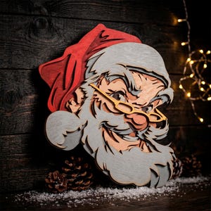 Può includere: Un'opera d'arte da parete in legno a strati che raffigura il volto di Babbo Natale. Il design presenta un cappello rosso, barba e baffi grigio chiaro e occhiali con montatura gialla. La costruzione multistrato crea profondità visiva.