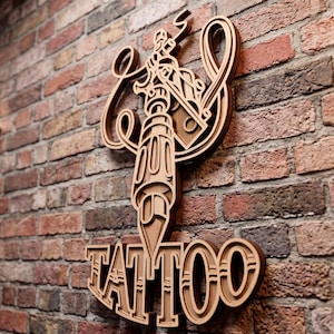 Può includere: Un'insegna da parete in legno a strati con un grafico stilizzato di una macchinetta per tatuaggi con intricate linee vorticose e la parola TATTOO in grassetto a blocchi alla base, caratterizzata da un design a strati di legno multidimensionale.