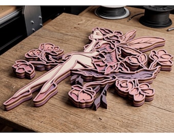 May include: A layered wooden fairy sculpture in shades of pink, purple, and beige. The fairy is seated among stylized flowers. The artwork is displayed on a wooden surface.