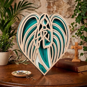 Op de afbeelding: Een gelaagd houten wandkunstwerk in de vorm van een hart met een gestileerde engelfiguur in het midden. Het ontwerp maakt gebruik van afwisselende tinten groenblauw, wit en natuurlijke houtkleuren om diepte te creëren. De engel heeft vleugels.