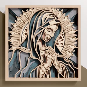 Puede incluir: Una obra de arte en papel en capas que representa una figura con un velo azul con las manos juntas en oración. La obra de arte presenta un halo y está enmarcada en un marco de madera de color claro. La paleta de colores incluye tonos de azul, beige y gris.