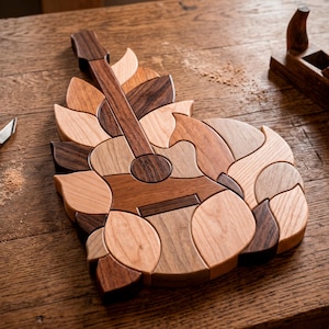 May include: A wooden guitar-shaped puzzle made from various wood tones, including light and dark browns. The puzzle is surrounded by leaf-shaped pieces, creating a decorative design. The image is taken on a wooden surface.