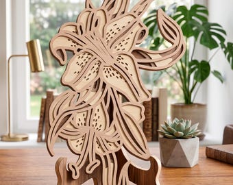 May include: A layered wooden lily sculpture in shades of light and dark brown. The floral design is intricately cut and stands on a wooden base. The sculpture is displayed on a wooden desk, with a window and plants in the background.