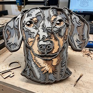 May include: A layered wooden dachshund dog head sculpture. The artwork features multiple layers of wood in shades of gray, brown, and tan, creating a 3D effect. The dog's eyes, nose, and ears are intricately detailed.