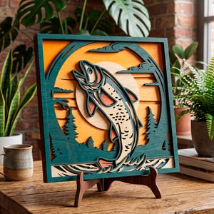 Op de afbeelding: Een gelaagd houten kunstwerk dat een springende vis tegen een zonsondergang afbeeldt. Het kunstwerk heeft een teal-kleurig frame en accenten, met de vis in beige, teal en koraal. Het wordt gepresenteerd op een houten ezel, ongeveer 30 cm vierkant.