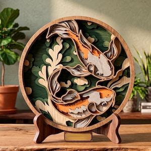 May include: A circular wooden artwork depicting two koi carp. The fish are orange, white, and grey, set against a green and beige backdrop. The layered design sits on a wooden stand, creating a 3D effect.