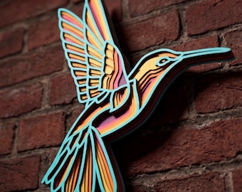 May include: A multi-layered wall art piece shaped like a hummingbird. The design features stacked layers in vibrant shades of turquoise, orange, yellow, and pink, creating a dimensional effect with visible cutouts that define the wings and tail feathers.