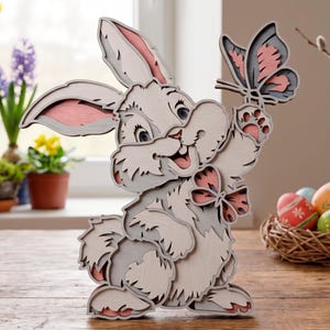 May include: A layered wooden cutout of a rabbit in shades of light gray and soft pink. The rabbit is depicted with a smiling expression, holding a butterfly in one paw, and features a small butterfly detail on its chest, creating a dimensional effect.