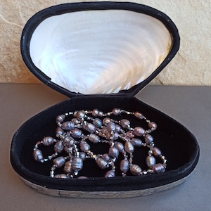 May include: A black velvet jewelry box shaped like a clam shell with a pearl-like interior. A strand of dark gray pearls and silver beads is inside the box.