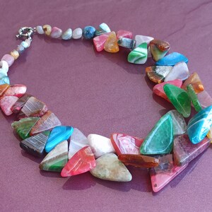 Pode incluir: Um colar colorido feito de pedras de ágata triangulares em vários tons de verde, azul, rosa, vermelho e marrom. O colar é enfiado em uma corrente de prata.