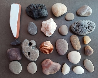 Può includere: Una collezione di pietre lisce grigie e marroni, conchiglie e alcuni pezzi di roccia vulcanica nera. Le pietre sono disposte su una superficie grigia.
