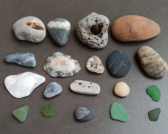 Può includere: Una collezione di pietre lisce e colorate, conchiglie e vetro di mare. Le pietre sono di varie tonalità di grigio, marrone e bianco. Le conchiglie sono bianche e hanno una lucentezza madreperlacea. Il vetro di mare è verde e blu.