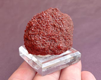 May include: A cluster of red crystals on a clear display stand. The crystals are small and have a rough texture.