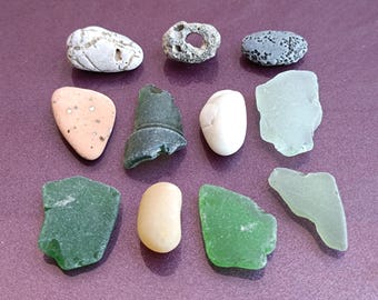 Può includere: Una collezione di dieci pezzi di vetro di mare e pietre da spiaggia in vari colori e forme, tra cui verde, blu, bianco, marrone e grigio. Le pietre sono disposte su una superficie viola.