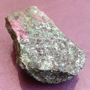 Peut inclure: Un spécimen de roche brute avec un mélange de couleurs. La pierre présente des zones de vert profond, parsemées de tons plus foncés, et un éclair de rose vif. La texture générale apparaît rugueuse et naturelle, suggérant un minéral ou une pierre précieuse.