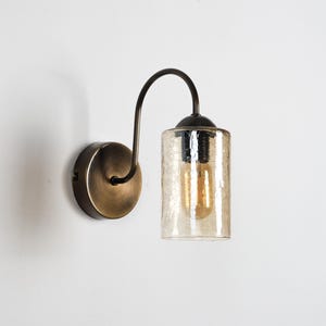 Peut inclure: Applique murale avec une finition bronze. Elle comprend un bras incurvé supportant un abat-jour cylindrique en verre texturé. L'ampoule à l'intérieur de l'abat-jour est visible, émettant une lueur chaude. Conçu pour une utilisation en intérieur.