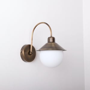 May include: A wall-mounted light fixture with a brass finish. The fixture features a curved arm, a conical shade, and a round, white globe. The design is reminiscent of vintage or industrial lighting.