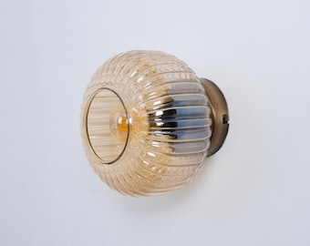 Könnte beinhalten: Eine goldene Wandleuchte mit einem gerippten, bernsteinfarbenen Glasschirm. Das Licht ist eingeschaltet und die Glühbirne ist sichtbar.