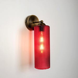 May include: A wall-mounted light fitting featuring a cylindrical red glass shade with a textured hobnail pattern. The lamp is attached to a circular, antique brass-finished metal base with a short, horizontal arm connecting to the light socket.