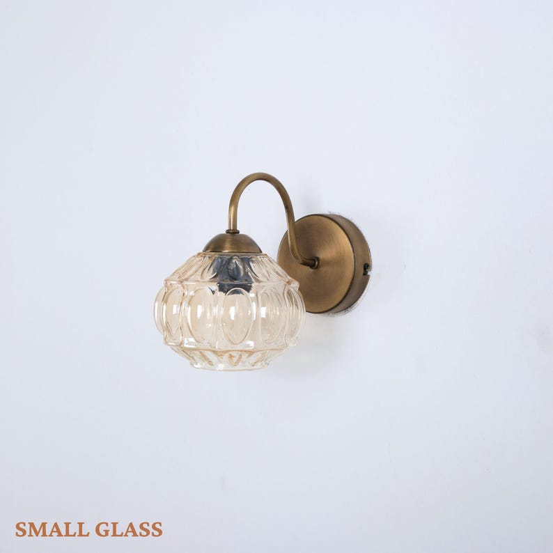 Puede incluir: Una l&aacute;mpara de pared de estilo vintage con una pantalla de cristal transparente estampada y un marco de metal color lat&oacute;n. La l&aacute;mpara tiene un brazo curvo y una base redonda para el montaje. El texto "SMALL GLASS" est&aacute; en la parte inferior de la imagen.