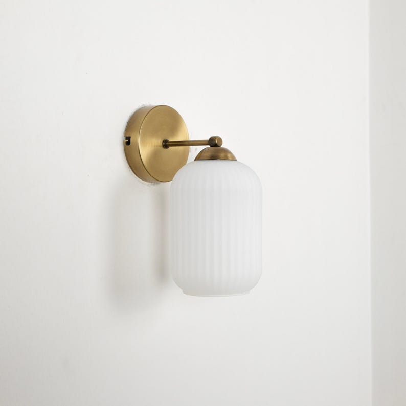 May include: A wall-mounted light fixture with a round, brass-colored base and a cylindrical, ribbed white glass shade. The fixture has a simple, elegant design, suitable for various interior styles. The light is attached to the wall with a brass arm.