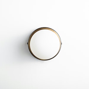 Op de afbeelding: Een ronde, zwarte wandlamp met een gouden rand en een witte, matte glazen kap.