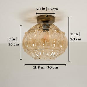 May include: A vintage-style ceiling light fixture. The fixture is 11 inches (28 cm) tall and 11.8 inches (30 cm) wide, with a 9-inch (23 cm) diameter. It features a bronze-colored base and a clear, amber-colored glass shade.