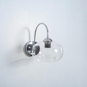 May include: A chrome wall sconce with a clear glass shade. The sconce has a curved arm and a round base.
