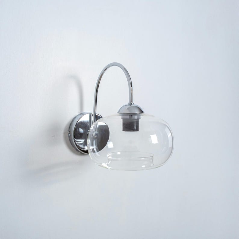 May include: A chrome wall sconce with a clear glass shade. The sconce has a curved arm and a round base.