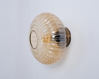 Könnte beinhalten: Eine goldene Wandleuchte mit einem gerippten, bernsteinfarbenen Glasschirm. Der Schirm ist rund und hat ein Loch in der Mitte für die Glühbirne.