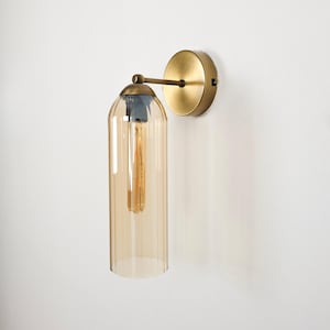May include: A wall-mounted light fixture with a brass finish. It features a cylindrical, amber-colored glass shade housing a vintage-style light bulb. The fixture includes a round backplate and a curved arm.