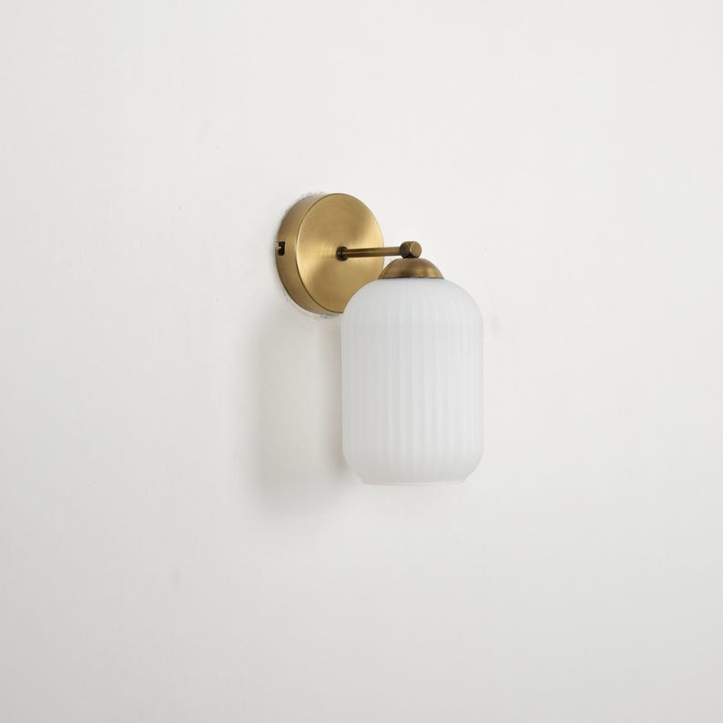 May include: A wall-mounted light fixture with a round, brass-colored base and a cylindrical, ribbed, white glass shade. The fixture has a simple, elegant design, suitable for various interior styles. The light fixture is mounted on a white wall.