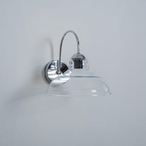 May include: A chrome wall sconce with a clear glass shade. The sconce has a curved arm and a round base.