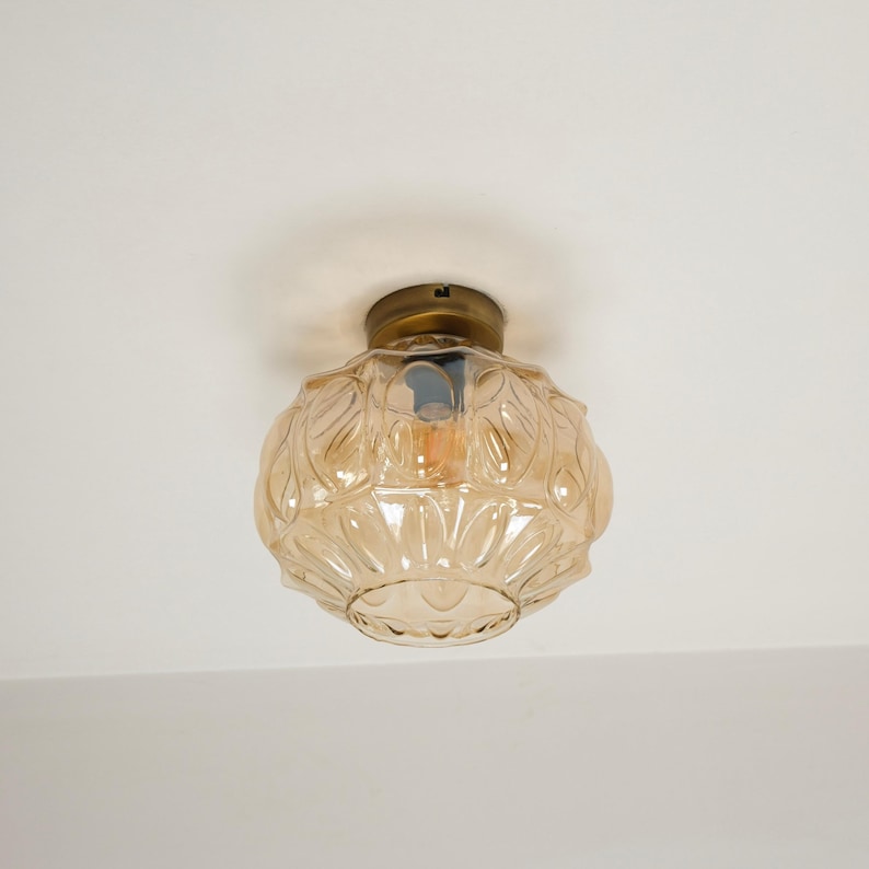 May include: A vintage-style ceiling light fixture with a decorative, amber-colored glass shade. The shade has a patterned design and is attached to a brass-colored base. The light bulb is visible inside the shade, casting a warm glow.