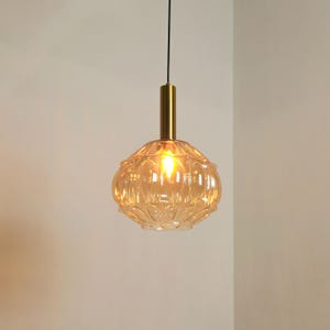 May include: A pendant light fixture featuring a spherical, amber-tinted glass shade with a textured, scalloped pattern. The shade is suspended by a thin black cord and attached to a cylindrical, brushed gold-tone metal hardware fitting at the top.