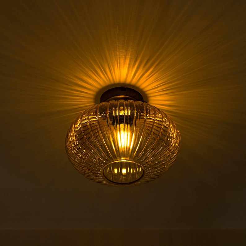 K&ouml;nnte beinhalten: Eine runde, bernsteinfarbene Glas-Deckenleuchte mit geripptem Design. Das Licht ist eingeschaltet und strahlt ein warmes Licht aus.
