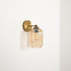 May include: A wall-mounted light fixture with a round, brass-colored base and a curved arm. The light has a ribbed, amber-colored glass shade. The fixture is designed for indoor use and adds a touch of vintage style.